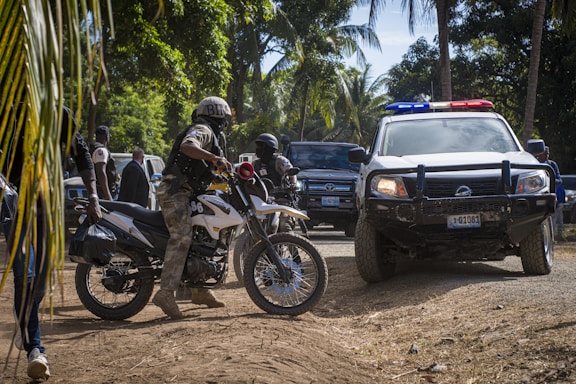 Several armed officers are situated around off-road vehicles in a wooded area. A motorbike with an officer is in the foreground, and a patrol vehicle with flashing lights is nearby. The scene suggests law enforcement activity in a tropical setting surrounded by palm trees and other greenery.