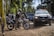 Several armed officers are situated around off-road vehicles in a wooded area. A motorbike with an officer is in the foreground, and a patrol vehicle with flashing lights is nearby. The scene suggests law enforcement activity in a tropical setting surrounded by palm trees and other greenery.