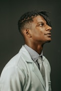 A person in a white lab coat is looking upwards with a thoughtful expression. They are wearing a checked shirt and a gray tie. Their hair is styled in twists and the background is plain and dark.