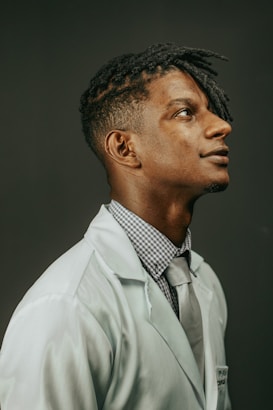 A person in a white lab coat is looking upwards with a thoughtful expression. They are wearing a checked shirt and a gray tie. Their hair is styled in twists and the background is plain and dark.