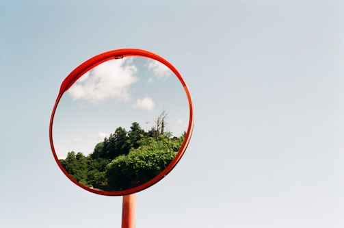 A convex safety mirror with a bright red rim reflects a lush green landscape and a partly cloudy sky, set against a plain blue sky in the background.