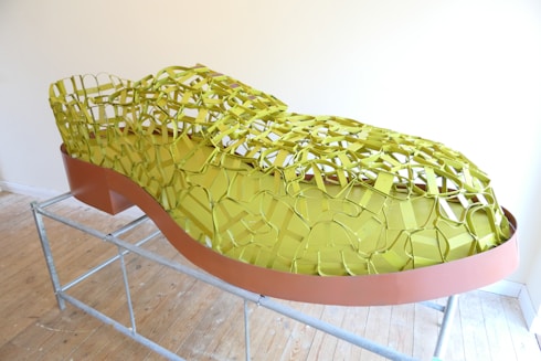 A large shoe sculpture constructed from metal mesh and solid components, displayed on a metal frame. The mesh part of the shoe is colored in a bright yellow-green, while the base is a reddish-brown. The sculpture rests on a wooden floor against a plain light-colored wall.