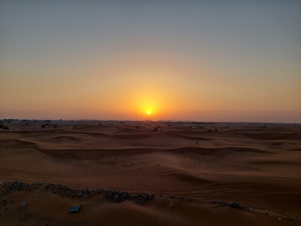 A vibrant sunset over rolling golden sand dunes with a 4x4 vehicle and camels in the distance.