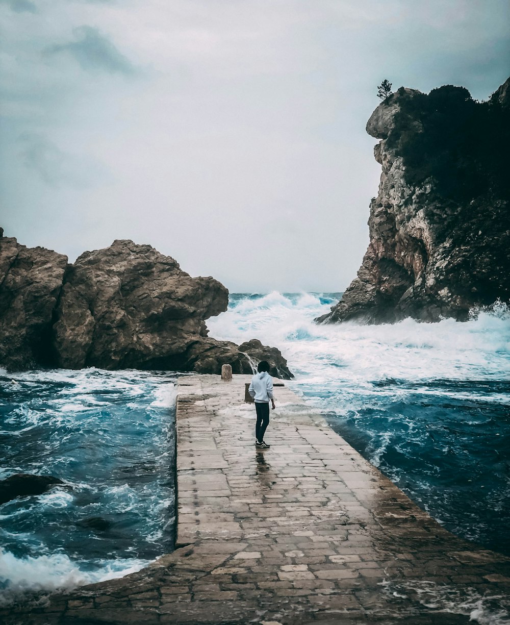 woman in white shirt and black pants walking on concrete pathway near sea during daytime