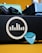 A close-up view of a music amplifier with a logo featuring vertical bars within a circle on its side. In front of it, a turquoise guitar pick is visible along with a black power cable partially connected. The background includes a teal and blue pedal unit, providing a colorful contrast to the composition.