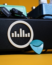 A close-up view of a music amplifier with a logo featuring vertical bars within a circle on its side. In front of it, a turquoise guitar pick is visible along with a black power cable partially connected. The background includes a teal and blue pedal unit, providing a colorful contrast to the composition.