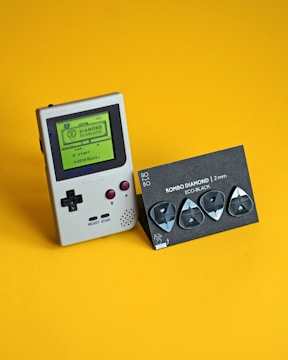 A vintage handheld gaming console with a green screen displaying text, positioned next to a set of black guitar picks on a yellow background. The console has maroon buttons and a directional pad, while the picks are attached to a labeled cardboard holder.