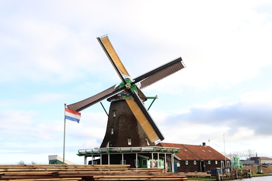 A traditional Dutch windmill with large sails is situated next to a small building with a red roof. The setting appears rural, with a clear blue sky and some clouds. A Netherlands flag is visible, adding a sense of national identity.