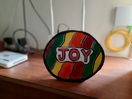 A multicolored, handcrafted sign displaying the word 'JOY' stands on a wooden surface. The sign features wavy stripes of red, yellow, orange, and green. In the background, there are various electronic devices and cords loosely organized against a plain wall.