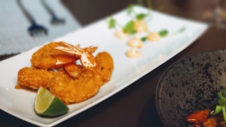 A white rectangular plate containing breaded and fried shrimp garnished with a lime wedge. In the background, there are small dollops of a creamy sauce and some green herbs on the plate. The image also shows part of a black plate with some food and green garnish on the right side and a small area with utensils in the upper left background.