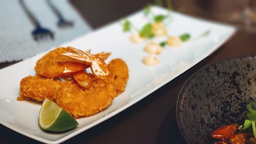 A white rectangular plate containing breaded and fried shrimp garnished with a lime wedge. In the background, there are small dollops of a creamy sauce and some green herbs on the plate. The image also shows part of a black plate with some food and green garnish on the right side and a small area with utensils in the upper left background.