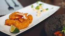 A white rectangular plate containing breaded and fried shrimp garnished with a lime wedge. In the background, there are small dollops of a creamy sauce and some green herbs on the plate. The image also shows part of a black plate with some food and green garnish on the right side and a small area with utensils in the upper left background.