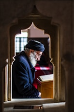 man in black suit jacket and black and white cap standing near brown wooden window