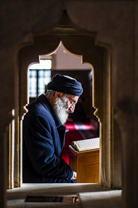 man in black suit jacket and black and white cap standing near brown wooden window