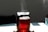 A glass kettle filled with steaming red liquid sits on a light-colored surface. Sunlight casts distinct shadows and highlights on both the kettle and the surface. A dark background contrasts with the lighter foreground, and a blue glass is partially visible in the background.