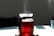 A glass kettle filled with steaming red liquid sits on a light-colored surface. Sunlight casts distinct shadows and highlights on both the kettle and the surface. A dark background contrasts with the lighter foreground, and a blue glass is partially visible in the background.