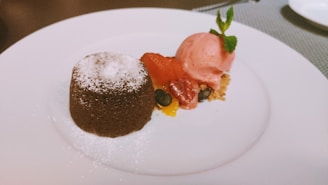 A chocolate lava cake dusted with powdered sugar sits on a plate next to a scoop of pink strawberry ice cream, garnished with fresh mint. Complementing the dessert are colorful sliced strawberries, blueberries, and small pieces of mango, placed on a layer of crumbled biscuit.