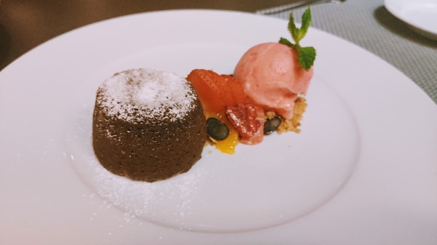 A chocolate lava cake dusted with powdered sugar sits on a plate next to a scoop of pink strawberry ice cream, garnished with fresh mint. Complementing the dessert are colorful sliced strawberries, blueberries, and small pieces of mango, placed on a layer of crumbled biscuit.