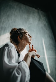 A woman is sitting in a contemplative pose, looking upward with a thoughtful expression. Her fingers are gently pointed towards her face, and she is wearing a white blouse with a dark underlayer. Soft lighting from the side casts subtle shadows.