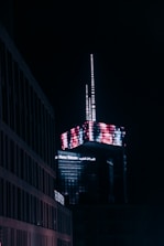 A modern skyscraper illuminated at night, featuring a complex arrangement of digital neon lights creating a vivid pattern. The building is branded with 'Maroc Telecom' signage. Surrounding structures appear dimly lit in comparison, emphasizing the vibrant top of the skyscraper.