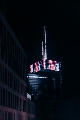 A modern skyscraper illuminated at night, featuring a complex arrangement of digital neon lights creating a vivid pattern. The building is branded with 'Maroc Telecom' signage. Surrounding structures appear dimly lit in comparison, emphasizing the vibrant top of the skyscraper.