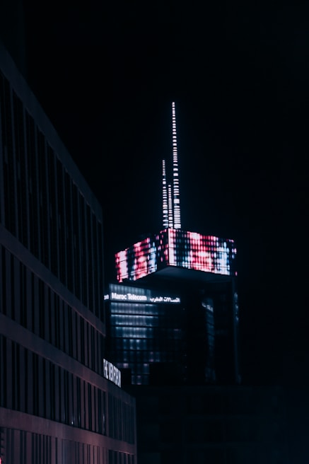 A modern skyscraper illuminated at night, featuring a complex arrangement of digital neon lights creating a vivid pattern. The building is branded with 'Maroc Telecom' signage. Surrounding structures appear dimly lit in comparison, emphasizing the vibrant top of the skyscraper.