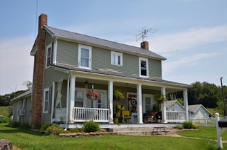 A two-story house with green siding and white trim. There is a large porch with white railing and several potted plants. Brick chimneys frame the structure, and an old-style TV antenna is on the roof. The house is surrounded by a well-kept lawn with a few trees and other small structures in the background.