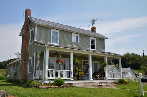 A two-story house with green siding and white trim. There is a large porch with white railing and several potted plants. Brick chimneys frame the structure, and an old-style TV antenna is on the roof. The house is surrounded by a well-kept lawn with a few trees and other small structures in the background.