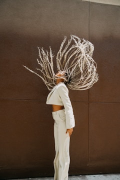 A person with braided hair is captured in motion, creating a dynamic silhouette against a plain, dark brown wall. Their outfit consists of a matching light-colored suit jacket and pants, revealing a confident and stylish demeanor.