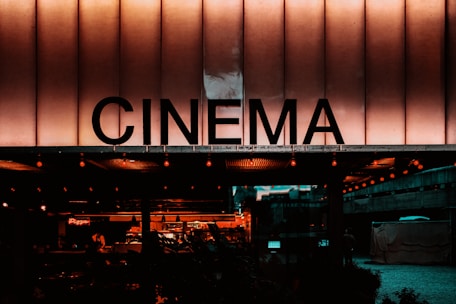 A building facade with a large illuminated sign displaying the word 'CINEMA' in bold letters. The scene has a warm, nostalgic glow with visible lights and a glimpse of an indoor area decorated with more lights and seating arrangements.