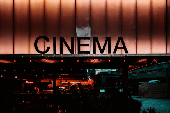 A building facade with a large illuminated sign displaying the word 'CINEMA' in bold letters. The scene has a warm, nostalgic glow with visible lights and a glimpse of an indoor area decorated with more lights and seating arrangements.