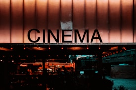 A building facade with a large illuminated sign displaying the word 'CINEMA' in bold letters. The scene has a warm, nostalgic glow with visible lights and a glimpse of an indoor area decorated with more lights and seating arrangements.
