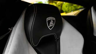 A luxurious car interior featuring a black leather seat with high-quality stitching and an embossed Lamborghini logo. The background shows blurred greenery, creating a contrast with the sleek, sophisticated design of the seat.