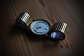 A gold wristwatch with a circular face lies on a wooden surface. The watch strap is metallic and flexible, with a ribbed design. The watch face displays Roman numerals and elegant, thin watch hands.