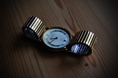 A gold wristwatch with a circular face lies on a wooden surface. The watch strap is metallic and flexible, with a ribbed design. The watch face displays Roman numerals and elegant, thin watch hands.