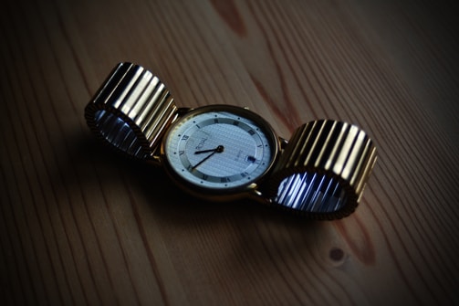 A gold wristwatch with a circular face lies on a wooden surface. The watch strap is metallic and flexible, with a ribbed design. The watch face displays Roman numerals and elegant, thin watch hands.