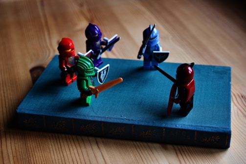 Several colorful toy figures resembling knights or warriors stand on a closed book. The figures have various colors like red, green, and blue, each holding swords and shields, suggesting a playful battle or gathering scene.