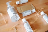 A wooden surface holds several clear plastic bottles with white labels, some with pump dispensers and others with twist caps. A rectangular metallic package lies among the bottles. Dried plant stems are scattered around, adding a natural element.