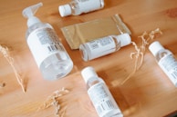 A wooden surface holds several clear plastic bottles with white labels, some with pump dispensers and others with twist caps. A rectangular metallic package lies among the bottles. Dried plant stems are scattered around, adding a natural element.