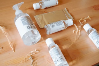 A wooden surface holds several clear plastic bottles with white labels, some with pump dispensers and others with twist caps. A rectangular metallic package lies among the bottles. Dried plant stems are scattered around, adding a natural element.