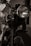 A close-up view of a vintage motorcycle, focusing on the front wheel and headlamp. The image is captured in black and white, highlighting the intricate details of the bike including the ribbed suspension forks and the gleaming metal surfaces. The background is softly blurred, emphasizing the motorcycle's classic design.