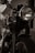 A close-up view of a vintage motorcycle, focusing on the front wheel and headlamp. The image is captured in black and white, highlighting the intricate details of the bike including the ribbed suspension forks and the gleaming metal surfaces. The background is softly blurred, emphasizing the motorcycle's classic design.