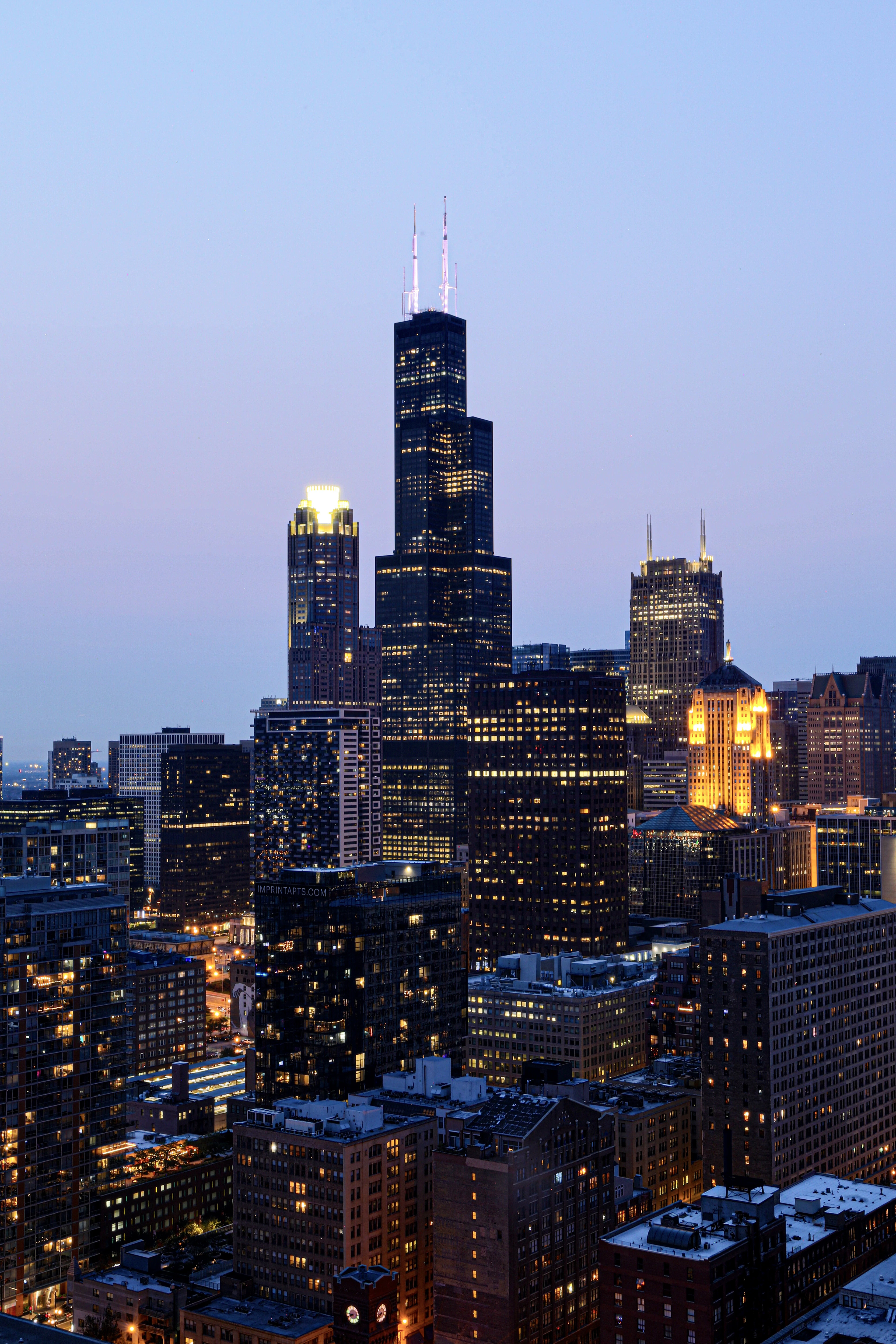 The Willis Tower's lit windows and antennas glow above the Chicago skyline at dusk.