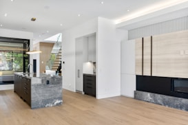 A modern kitchen with sleek, minimalist design featuring a large island with a marble countertop and built-in sink. The room has wooden flooring and neutral-colored cabinets, with a staircase leading to an upper floor. Large windows allow natural light to fill the space, enhancing its open and airy feel.