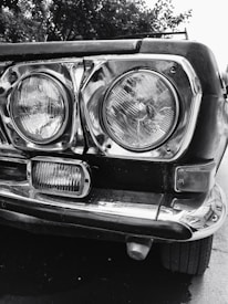 Close-up view of the front part of a vintage car, showcasing two large, round headlights encased in chrome and a smaller rectangular light below. The metal frame is slightly worn, adding character to the vehicle. In the background, there are blurry trees.