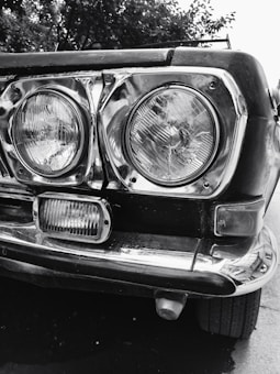 Close-up view of the front part of a vintage car, showcasing two large, round headlights encased in chrome and a smaller rectangular light below. The metal frame is slightly worn, adding character to the vehicle. In the background, there are blurry trees.