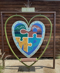 A large heart-shaped frame is set against a wooden backdrop. Within the frame, a colorful puzzle piece artwork depicting mountains, forests, and water is visible. A small bench is positioned under the heart, and a sign with 'Love Forever' is placed above the artwork.