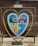 A large heart-shaped frame is set against a wooden backdrop. Within the frame, a colorful puzzle piece artwork depicting mountains, forests, and water is visible. A small bench is positioned under the heart, and a sign with 'Love Forever' is placed above the artwork.