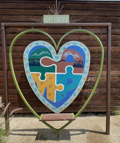A large heart-shaped frame is set against a wooden backdrop. Within the frame, a colorful puzzle piece artwork depicting mountains, forests, and water is visible. A small bench is positioned under the heart, and a sign with 'Love Forever' is placed above the artwork.