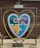 A large heart-shaped frame is set against a wooden backdrop. Within the frame, a colorful puzzle piece artwork depicting mountains, forests, and water is visible. A small bench is positioned under the heart, and a sign with 'Love Forever' is placed above the artwork.